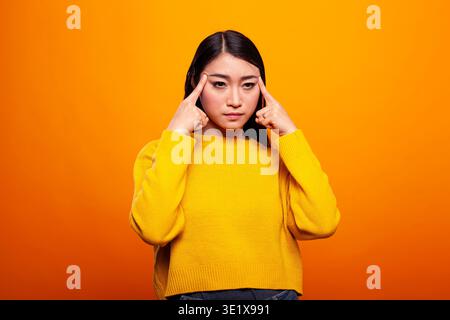 Asiatische weibliche Person, die die Schläfe berührt, tiefe Gedanken mit einem ernsthaften Ausdruck im Gesicht hat. Porträt einer Frau mit intensivem Blick, die mit ihren Zeigefingern die Seiten ihres Kopfes drückt. Stockfoto