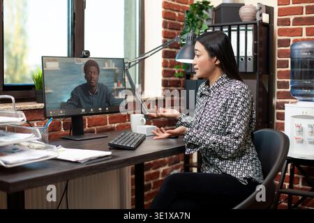 Unternehmerin, die virtuelles Meeting mit männlichen Kunden am Schreibtisch führt und Marketingstrategien mithilfe des Computers weitergibt. Asiatischer Mitarbeiter in einem Büro, der mit einem Remote Manager im Videoanruf spricht. Stockfoto