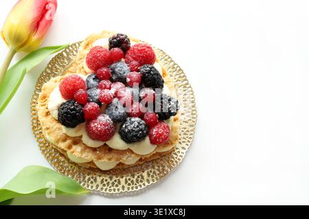 Valentinstag. Köstliche herzförmige Fruchttorte mit frischen Beeren und Puderzucker, rote Tulpen auf weißem Hintergrund. Draufsicht. Stockfoto