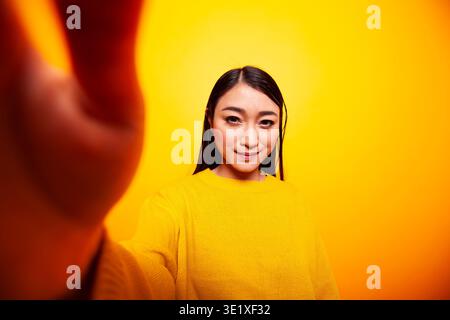 Lächelnde asiatische Einzelperson, die die Kamera hält und ein Video für ihr Online-Publikum macht. Schöne Frau, die ein Selfie vor einem isolierten gelben Hintergrund macht, pov-Konzept. Stockfoto