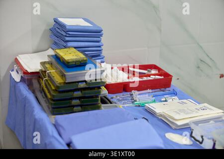 OP-Vorbereitungstisch mit gestapelten sterilen Instrumententabletts, Spritzen, Fläschchen, Gaze, Vorhängen und medizinischem Zubehör auf blauer steriler Abdeckung. Stockfoto