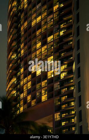 Nächtliche Fotografie einer modernen Wolkenkratzerfassade mit Reihen beleuchteter Fenster und Balkonen mit gelbem Innenlicht in der Stadt. Stockfoto