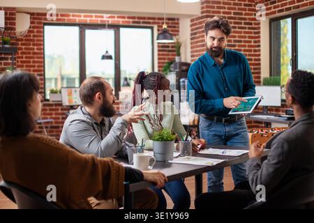 Weißbärtiger Mann, der mit einem Tablet steht und Projektaktualisierungen mit Kollegen in modernen Büros teilt. Das vielfältige Startup-Team hört den männlichen Analysten aufmerksam zu, die bei Gruppenbesprechungen finanzielle Präsentationen machen Stockfoto