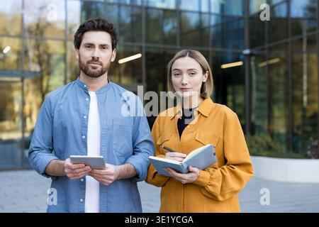 Geschäftspartner, die im Freien in der Nähe eines modernen Bürogebäudes stehen, ein Mann in der Hand eines digitalen Tablets und eine Frau, die ein offenes Notebook mit einem Stift hält und beide direkt in die Kamera blicken Stockfoto