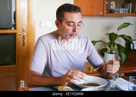 Mann mittleren Alters, der in heller Küche zwanglos isst und sein Smartphone überprüft Stockfoto