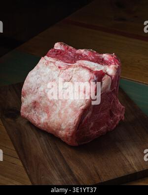 Roher Rindfleischbraten liegt auf dunklem Holzschneidebrett. Reichhaltiges rotes Fleisch mit marmoriertem Fett und Knochen. Holzmaserung zeigt die Textur unter dem Braten. Warme Beleuchtung Hi Stockfoto