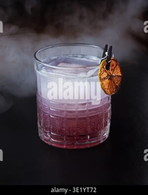 Rauchiger Cocktail in strukturiertem Glas mit einer getrockneten Orangenscheibe. Rosafarbene und klare flüssige Schichten stehen im Kontrast zu einem dunklen Hintergrund. Frostige Eiswürfel kühlen Stockfoto