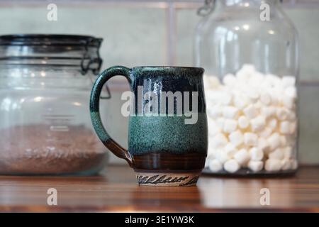 Handgemachte grüne Tasse mit Schokoladenpulver und Marshmallows in Glasbehältern im Hintergrund, um ein leckeres heißes Schokoladengetränk zu machen Stockfoto