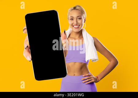 Frau zeigt das Smartphone auf hellem Hintergrund, während sie lächelt und Fitnesskleidung trägt Stockfoto