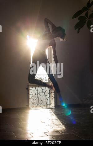Elegante Silhouette einer Frau, die mitten im Sprung in einer anmutigen, tänzerartigen Pose festgehalten wurde und mit einem Bein zum Knie hin balanciert. Hintergrundbeleuchtung durch Fenster Stockfoto