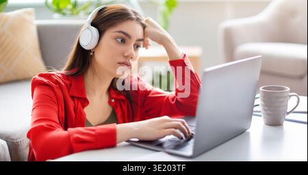 Die junge Frau konzentrierte sich auf ihren Laptop, während sie Kopfhörer in einem hellen Raum trug Stockfoto