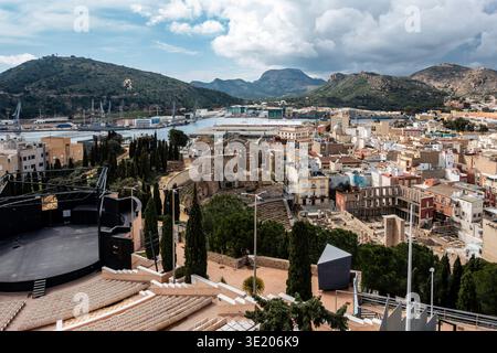 Ein weitläufiger Blick in Cartagena, Spanien, zeigt die Ruinen des römischen Theaters, ein Auditorium im Freien und den Industriehafen vor Bergen. Stockfoto