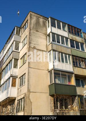 Apartmentblock im altehrwürdigen sowjetischen Stil mit geschlossenen Balkonen und verwitterter Fassade unter blauem Himmel mit typisch osteuropäischen Häusern Stockfoto