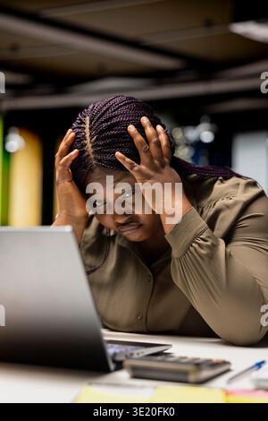 Schwarze Assistentin verspannt und belastet im dunklen Büro nach Stunden, erlebt Herausforderungen und Ängste am Arbeitsplatz. Entmutigte Frau, die mit intensiver Arbeit und ohne Freizeit zu kämpfen hat. Stockfoto