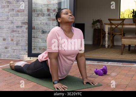 Afroamerikanerin, die Kobra auf grüner Matte spielt, Backsteinterrasse, in Leggings, Kurzhanteln Stockfoto