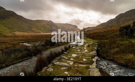 Moody Mountain Valley Landschaft mit Steinpfad, der durch feuchte Moore in Richtung entfernter See führt, unter dramatischem bewölktem Himmel bei Sonnenuntergang. Stockfoto
