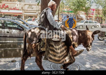 YAZD, IRAN - 18. AUGUST 2016: Ein älterer Mann reitet auf einer Straße in Yazd, Iran, auf einem Esel, der sich durch modernen Verkehr und Ladenfronten bewegt und die Tradition widerspiegelt Stockfoto