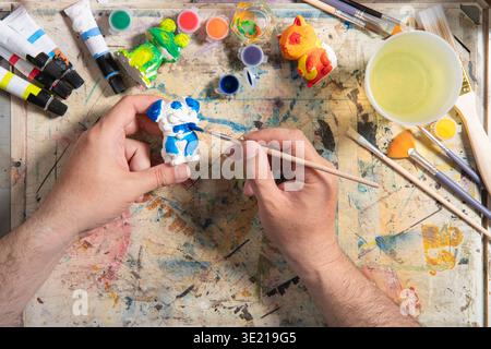 Draufsicht auf die Hände eines Mannes in einem blauen Hemd, der einen feinen Pinsel mit orangefarbener Farbe hält und eine kleine weiße Keramikfigur auf einem Arbeitstisch mit Farbglas malt Stockfoto