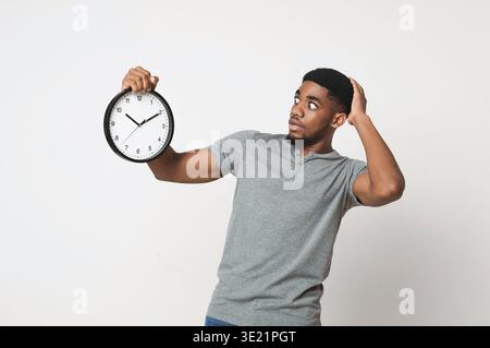 Schockierter schwarzer Mann, der eine Wanduhr auf weißem Hintergrund hält Stockfoto