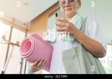 Aktive reife asiatische Frau im Fitnessstudio, SMS auf dem Smartphone und trage Yoga-Matte während der morgendlichen Fitness-Routine. Selektiver Fokus. Stockfoto