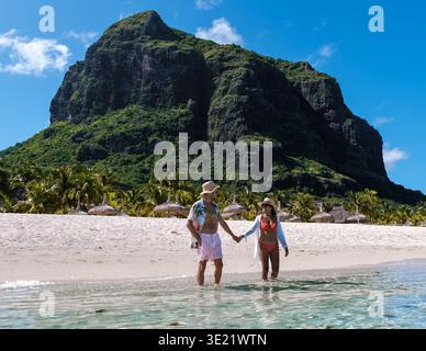 Ein glückseliges Paar schlendert Hand in Hand durch das klare türkisfarbene Wasser eines Strandes auf Mauritius. Hoch aufragende Klippen und pulsierende Palmen bilden eine atemberaubende Kulisse für ihr Urlaubsabenteuer. Stockfoto