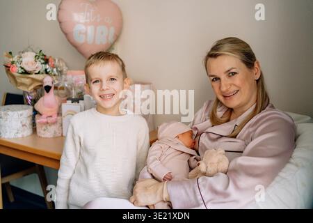 Mutter, die das Neugeborene hält, während sie mit dem älteren Sohn zu Hause steht. Wunderschönes Familienporträt, das Liebe, Eltern und Geschwisterverbindung zeigt. Stockfoto