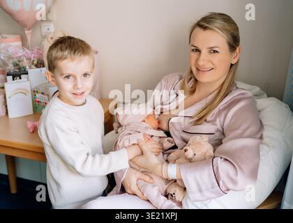 Mutter hält das Neugeborene, während das ältere Kind neben ihr steht. Vorstellung von Mutterschaft, Elternschaft und glücklichem Familienleben. Stockfoto