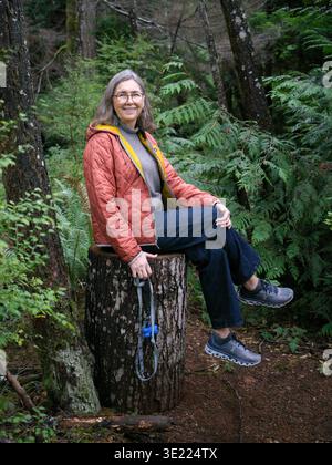 Frau in lässiger Outdoor-Kleidung sitzt auf einem Baumstumpf in einer üppigen Waldlandschaft. Bowser, Vancouver Island, Kanada Stockfoto