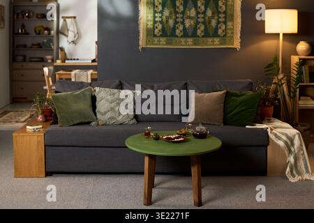 Wohnzimmerszene mit grünem runder Couchtisch mit Tee-Set und Snacks vor grauem Sofa mit mehreren Kissen, Hintergrund mit Bücherregal, Lampe und dekorativem Wandaufhängen Stockfoto
