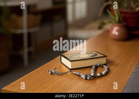 Islamische Gebetskegeln ruhen neben dem geschlossenen Koran auf Holztisch, im Vordergrund stehen religiöse Objekte mit verschwommenem Hintergrund, was auf eine friedliche Innenumgebung hindeutet Stockfoto