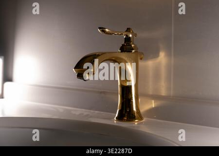 Goldener Wasserhahn glänzt neben dem weißen Waschbecken. Weiches Licht wirft sanften Schatten auf gewölbte Oberflächen. Modernes Design mit elegantem, minimalistischem Griff. Pur Stockfoto