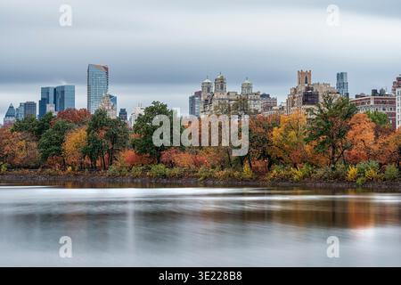 Skyline der Stadt mit Herbstbäumen, die sich in einem ruhigen See unter einem bewölkten Himmel spiegeln. Central Park Lake, New York City, USA Stockfoto