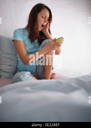 Frau, die auf dem Bett sitzt, gähnt, während sie den Wecker hält und blauen Pyjama trägt. Deutschland Stockfoto