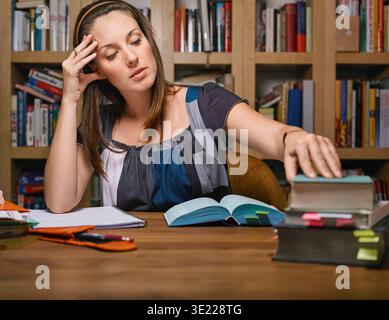 Frau, die an einem Holztisch mit Büchern und Notizen in einer gemütlichen Bibliothek studiert. Deutschland Stockfoto
