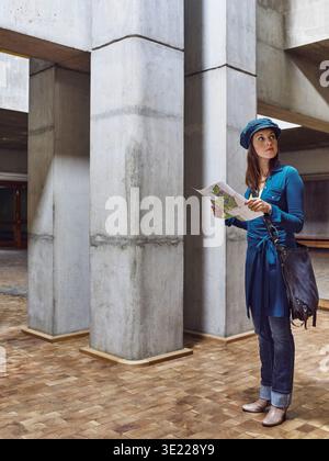 Eine Frau in lässiger Kleidung, die eine Karte hält, steht in einem modernen Betongebäude. Deutschland Stockfoto