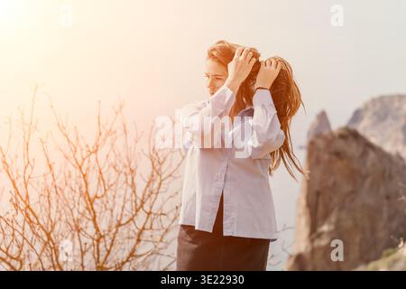 Reisefrau Natur Junge Frau, die ihr langes Haar während der goldenen Stunde auf einer Bergklippe mit malerischem Blick auf das Meer fixiert Stockfoto
