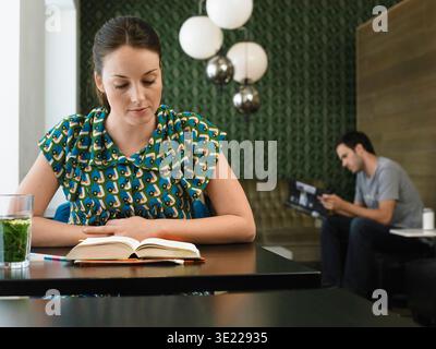 Frau liest ein Buch in einem Café, während ein Mann im Hintergrund eine Zeitschrift liest. Deutschland Stockfoto