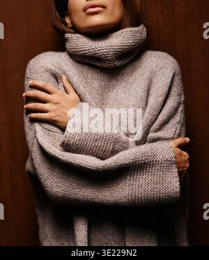 Frau in einem kuscheligen beigen Strickpullover, der sich vor einem hölzernen Hintergrund umarmt. Deutschland Stockfoto