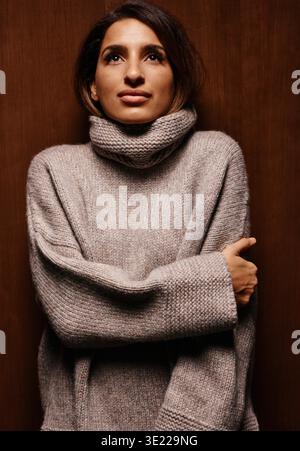 Frau in einem gemütlichen Rollkragenpullover steht vor einem hölzernen Hintergrund und kreuzt ihre Arme. Deutschland Stockfoto