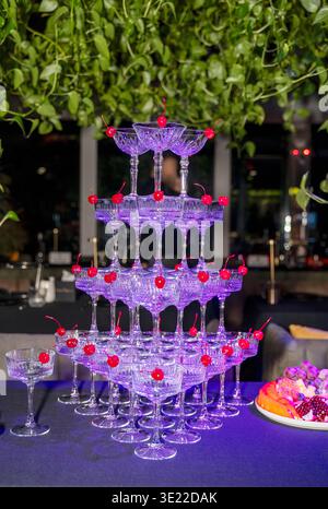 Champagnerturm mit roten Kirschen in jedem Glas unter violetter Beleuchtung. Konzept von professionellem Catering-Service, festlicher Feiertagsgala und Raffinesse Stockfoto