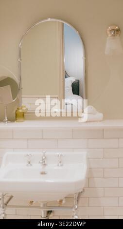Sanftes Licht taucht in ein altes Waschbecken im Badezimmer. Der geschwungene Spiegel spiegelt die gemütliche Schlafzimmerszene wider. Weiße Fliesen umrahmen ein klassisches Standwaschbecken. Das Handtuch liegt ordentlich auf Stockfoto