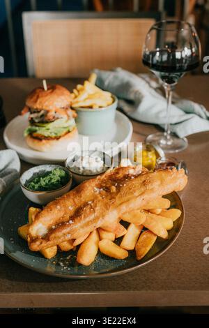 Goldenes Fischfilet liegt auf knusprigen Pommes Frites, einladend überzogen. Frisch gebackener Burger steht neben einer kleinen Schüssel Chips. Ein Glas Rotwein rundet das c Stockfoto