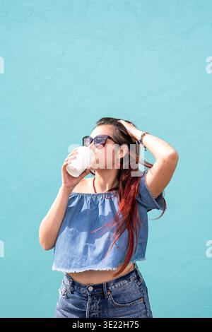 Attraktive junge Frau in Sommerkleidung und Sonnenbrille, die eine Tasse Kaffee in den Händen hält, auf blauem Hintergrund auf der Straße tanzt || Model veröffentlicht Stockfoto
