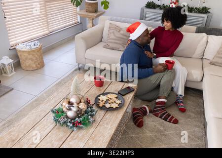 Afroamerikanisches Paar, das auf einem Sofa im Wohnzimmer sitzt und Kekse und rote Tassen teilt. Gemütlich, festlich, Urlaub, Dekor, Herzstück, Greenery, Kugeln || Modell veröffentlicht Stockfoto