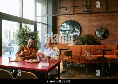 Junger Mann und Frau teilen sich Pizza und lachen im modernen Café Stockfoto