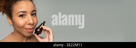 Eine schöne junge Frau hält ein Hautpflegeprodukt und lächelt strahlend vor einem minimalen Hintergrund. Stockfoto