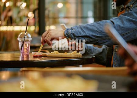 Junger Erwachsener Mann, Der Das Smartphone Benutzt, Während Er Drinnen Pommes Frites Isst Stockfoto