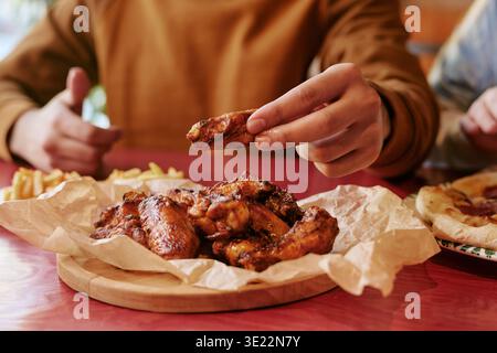 Junger Mann isst Chicken Wings am Restauranttisch mit Freunden Stockfoto