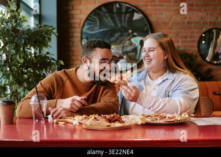 Junge Frau Füttert Jungen Mann Aus Dem Nahen Osten Pizza, Während Sie Drinnen Lächelt Stockfoto