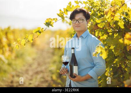 Gut aussehender asiatischer Ethnicity Sommelier mit geöffneter Flasche Rotwein und gegossenem Glas auf dem Weinberg Stockfoto